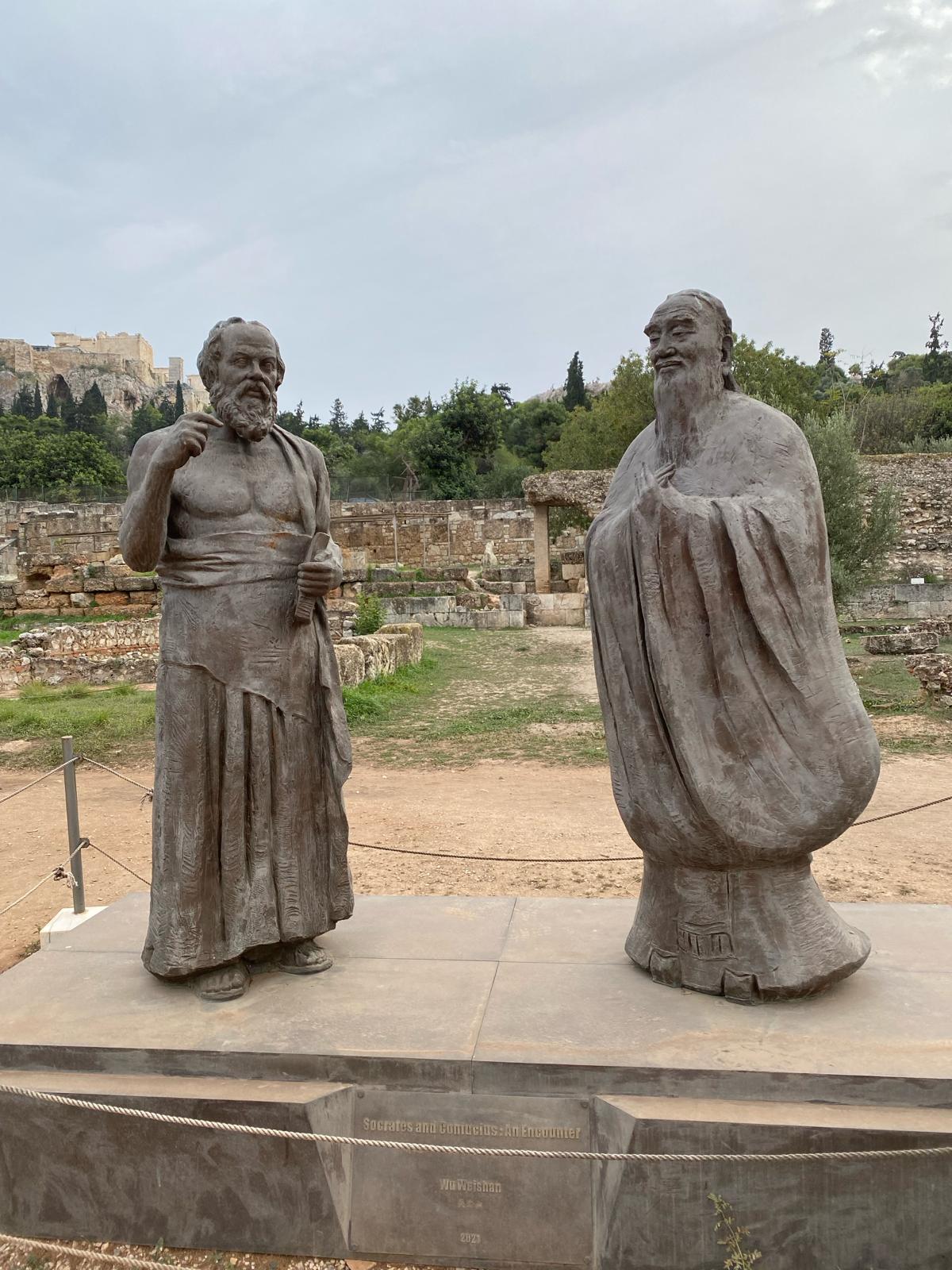 Zwei Bronzestatuen der Philosophen Sokrates (links) und Konfuzius (rechts) in einem Park mit archäologischen Ausgrabungen. Im Hintergrund das Pantheon in Athen.